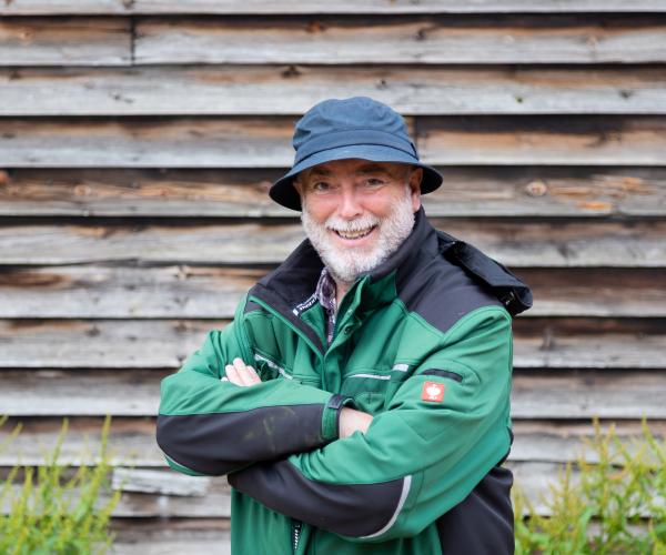 Phil standing with his arms crossed in front of a wooden panelled wall wearing a black bucket hat and a green branded jacket.