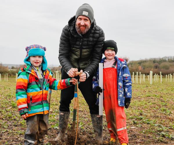 Father and two young sons stood smiling in a tree planting field holding a spade