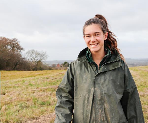 Forest Ranger Phoebe standing in a field smiling with her arm resting on a wooden tree stake, wearing a dark green waterproof coat, with newly planted trees in tubes behind her.