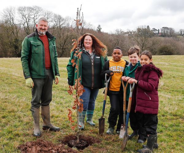 Head Forester Stephen, Chief Executive Beth Brook, and three school children all holding spades stand around an English oak tree sapling in a hole in the ground. They are all looking at the camera and smiling.
