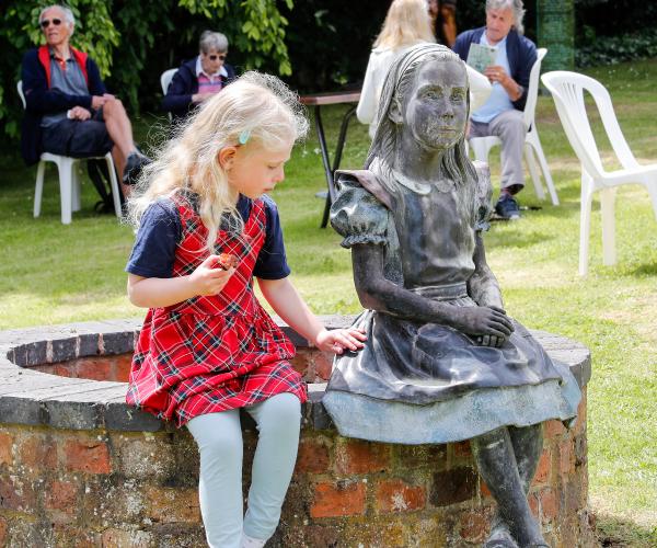 A young blonde girl in a red dress sat next to a bronze sculpture of Alice, from Alice in Wonderland.