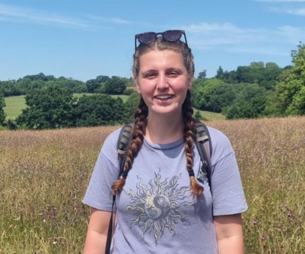 Becky, Biodiversity Intern, standing in a field with trees in the background. 