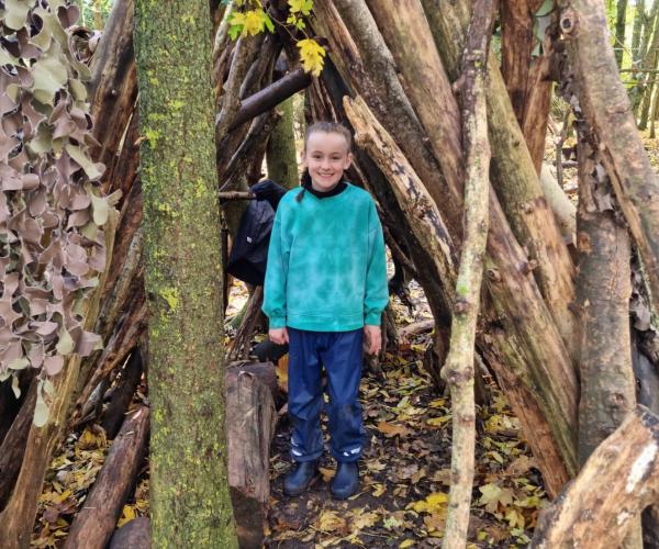 A young girl standing in a den made of tree branches in a woodland clearing smiling at the camera