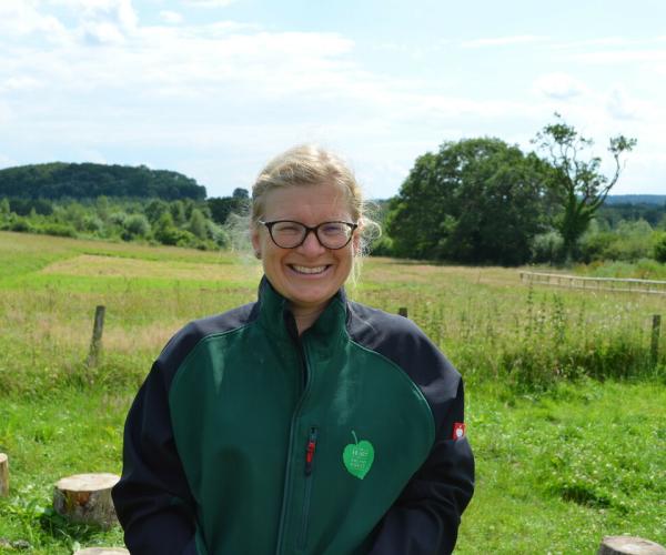 Andrea wearing the Heart of England Forest branded clothing, smiling at the camera with windmill hill in the background