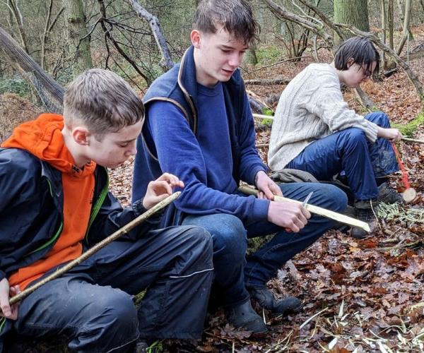 Three young people sat along a log in a forest clearing whittling during a Young Foresters club session