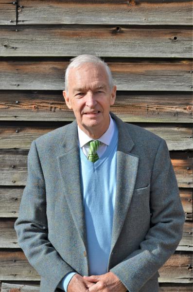 Jon Snow standing in front of a wooden panelled wall wearing a suit and tie. 