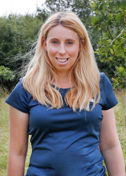 Close up photo of Ellie, our Gorcott Outdoor Learning Officer, stood smiling in front of Forest background wearing a bright blue t-shirt.