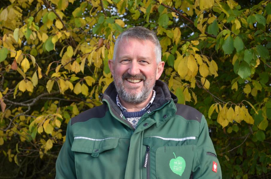 Head and shoulders photo of Head Forester Stephen wearing a Heart of England Forest branded jacket standing in front a tree with yellow autumn leaves