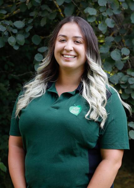 Close up portrait of Tasha, our Gorcott Biodiversity Officer, stood smiling in front of green leaves, wearing a green, branded polo shirt.