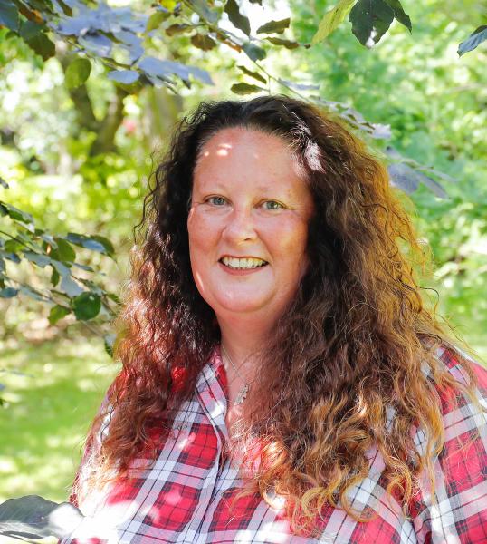 Head and shoulders photo of Beth Brook stood under a tree with green leaves on a sunny day