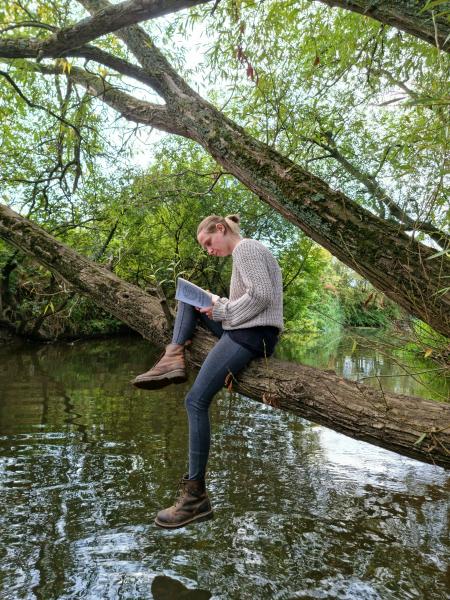 Our Marketing and Communications Officer, Julie, sitting on a branch over the river reading a book