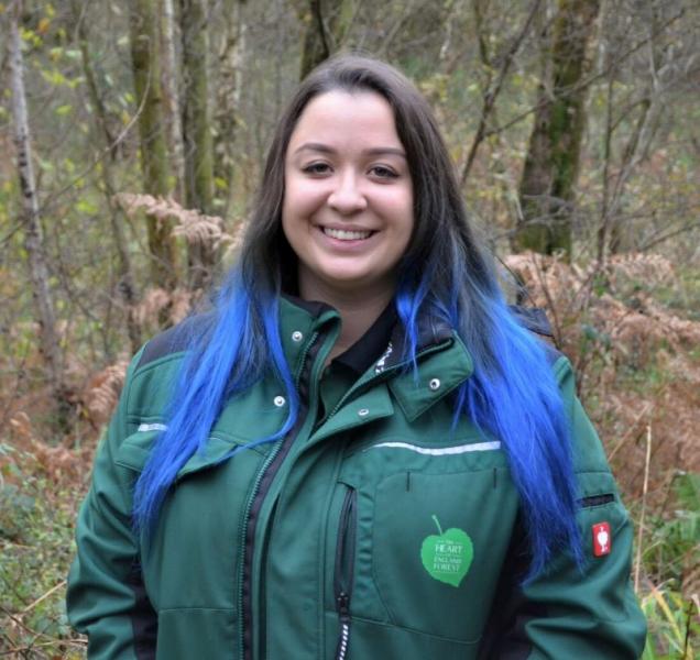 Tasha, our Gorcott Biodiversity Officer, is standing in the woodland, looking and smiling at the camera wearing a Heart of England Forest branded coat 