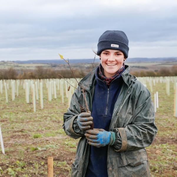Assistant Forest Ranger Melissa standing in a field with newly planted trees behind her. She is wearing warm clothes and a hat, holding a tree sapling and smiling at the camera