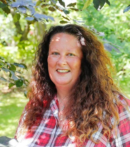 Beth Brook, Chief Executive, Heart of England Forest, standing and smiling at the camera with tree branches and green leaves above her