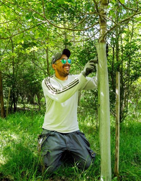 Leon, a volunteer, crouching down and cutting a protective tree guard off a young established tree in the Forest