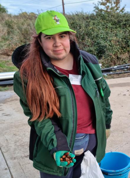 Rhiannon, our Tree Nursery Assistant, standing holding a handful of acorns. She is wearing a lime green cap and a green branded Heart of England Forest coat