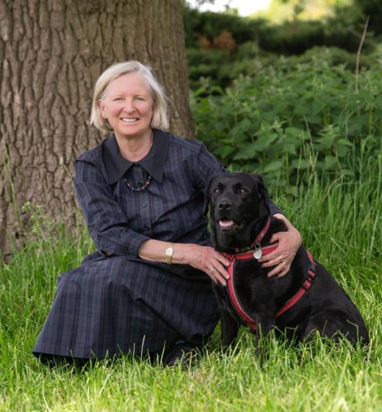 Heather Acton taken crouching down on the grass by a large tree trunk with her black Labrador sitting besides her