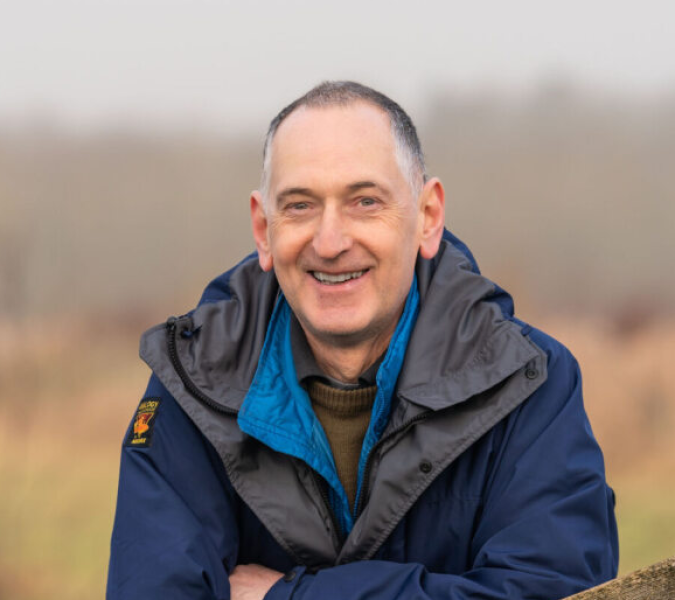 Rob Mackenzie, leaning on a post. He is smiling at the camera. This has been captured as a head and shoulders shot. 