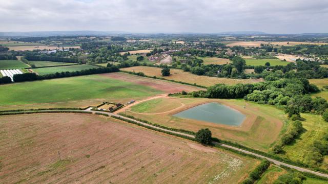 Aerial view of a small reservoir at Luddington, surrounded by fields, hedgerows and patches of trees, with open countryside stretching into the distance under a cloudy sky.