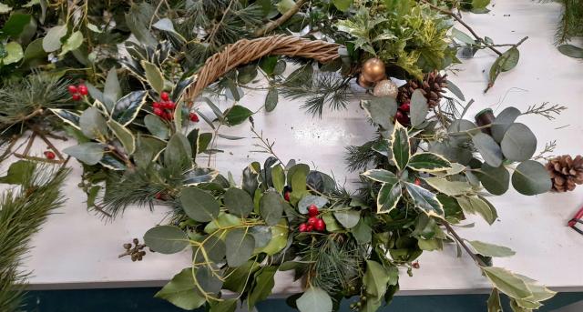 A handmade Christmas wreath decorated with eucalyptus, holly, pine sprigs, red berries, and pinecones on a table covered with greenery.