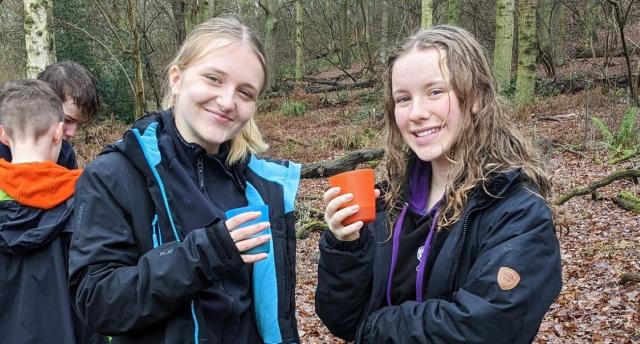 Two young people smiling at the camera and enjoying a hot drink during a woodland activity in the Heart of England Forest.